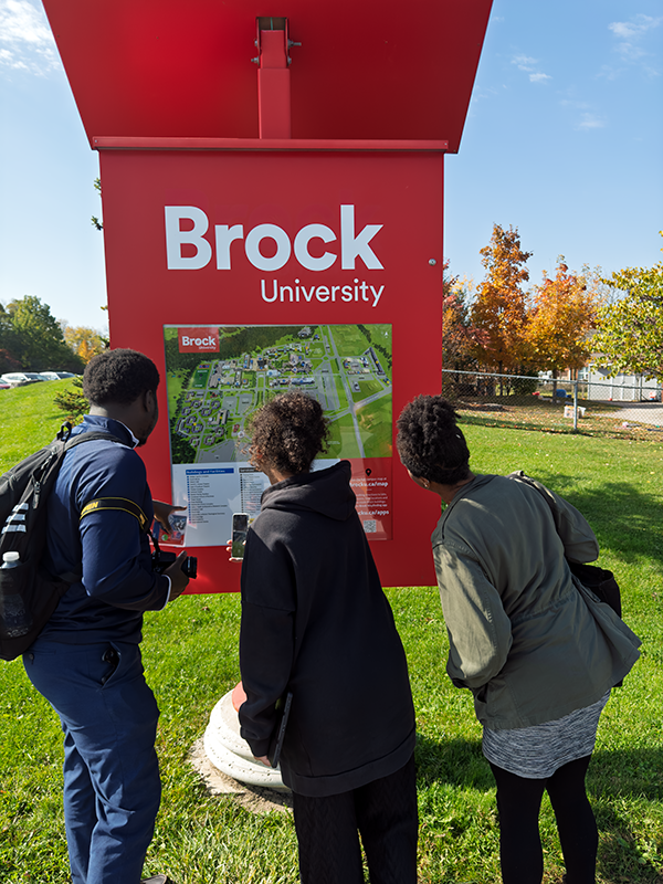 Alex Allasra, Gabriela Sealy, and Dr. Émilie Andrée Roumer Jabouin look at a large red orientation map at Brock University. Autumn trees and a bright sky appear in the background. Photo by Blessing Ogunyemi.