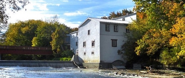 White building with a sloping roof. A wooden bridge spans the river dike in the foreground.