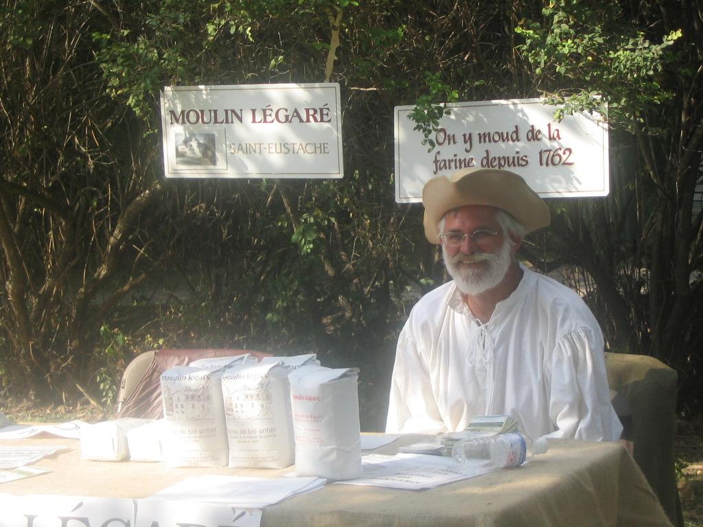 Pierre-Paul Renaud, sporting a white beard, a beige three-cornered hat and a white shirt, sits outside the mill behind a flour sale table.