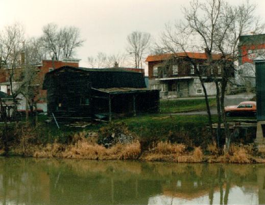 Wide shot of a darkened, weathered building next to a river.