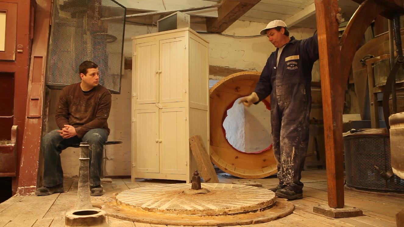 Wide shot of Martin Trudel sitting on the left, looking at Daniel Saint-Pierre standing on the right side of the photo, dressing the lower millstone.