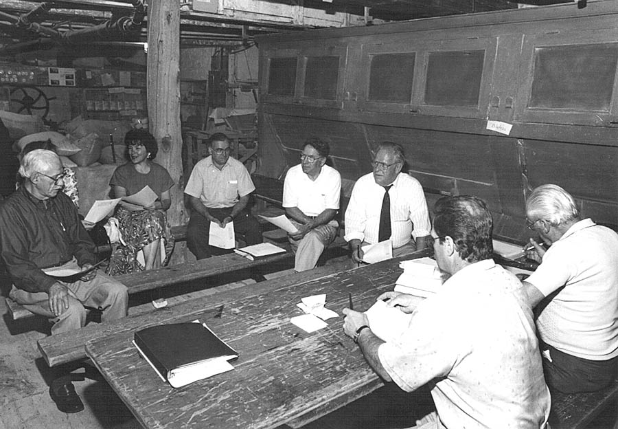 Black and white photo of seven board members sitting in a circle on wooden benches, holding sheets of paper.