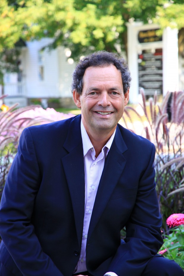 Medium close-up of Pierre Charron with short brown curly hair, wearing a navy jacket and striped shirt, sitting outside in front of some flowers.