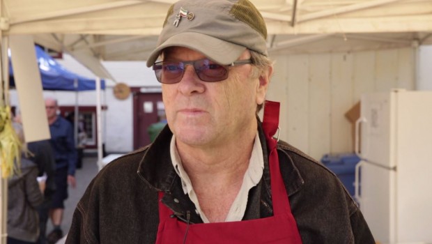 Close-up of Luc Raby wearing sunglasses, a khaki cap and a red apron.