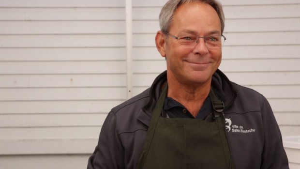 Close-up of Patrice Paquette with grey hair and glasses, wearing a green apron, and smiling at the camera.