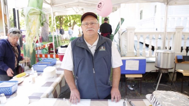 Waist-up shot of Raymond Tessier, with glasses, a cap and a blue jacket, standing under a tent.