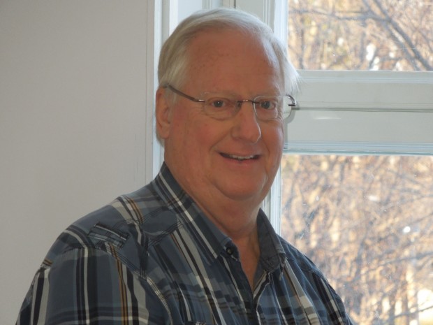 Close-up of Roger Pomerleau with white hair, wearing a plaid shirt, standing in front of a window.