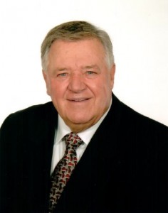 Bust shot of Guy Bélisle with short grey hair, wearing a black jacket and a tie.