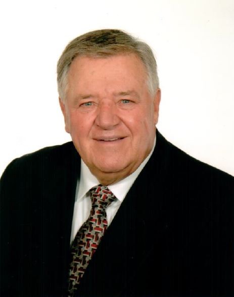 Bust shot of Guy Bélisle with short grey hair, wearing a black jacket and a tie.