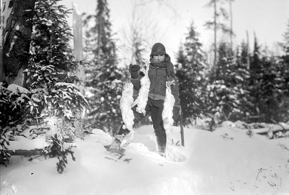 Black and white picture of a young kid in snowshoes with three rabbits in hand, taken by hunting in the forest.