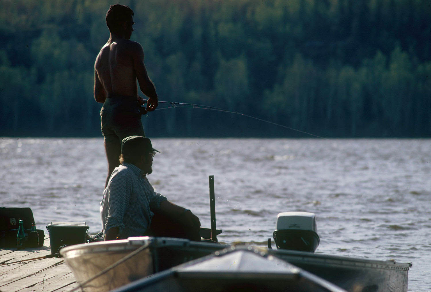 Photographie couleur de deux hommes pêchant sur un quai devant un lac.
