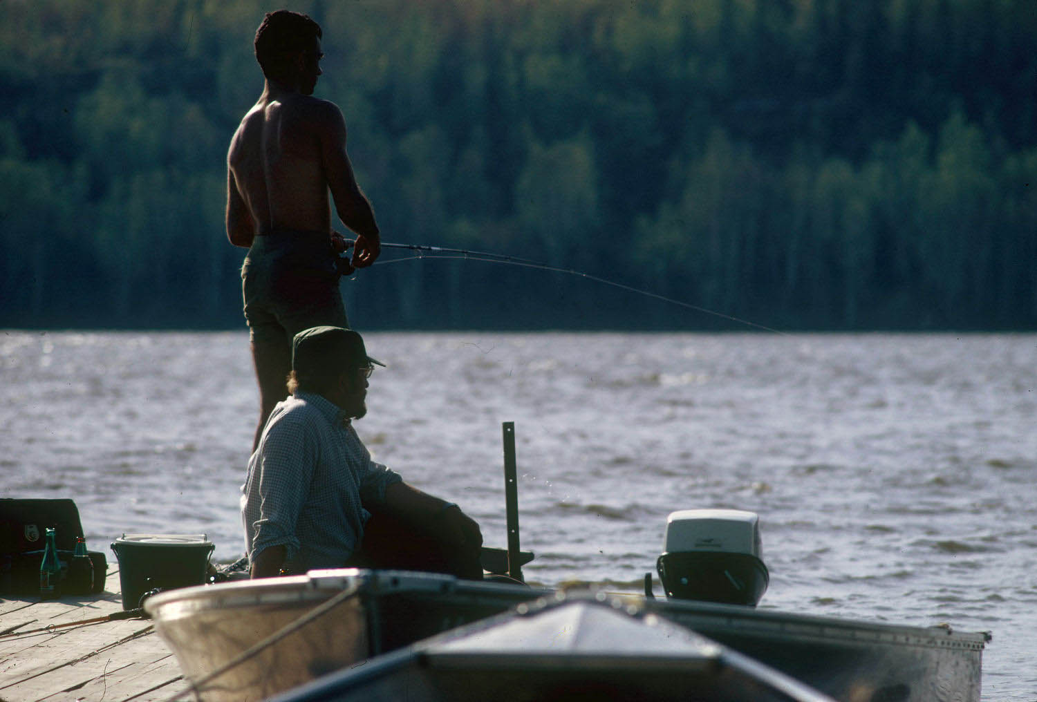 Coloured picture of two men fishing on a dock in front of a lake.