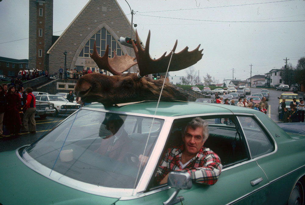Coloured picture of a car with a moose head fixed at the top during a parade around the city. A man behind the wheel looking at the photographer.