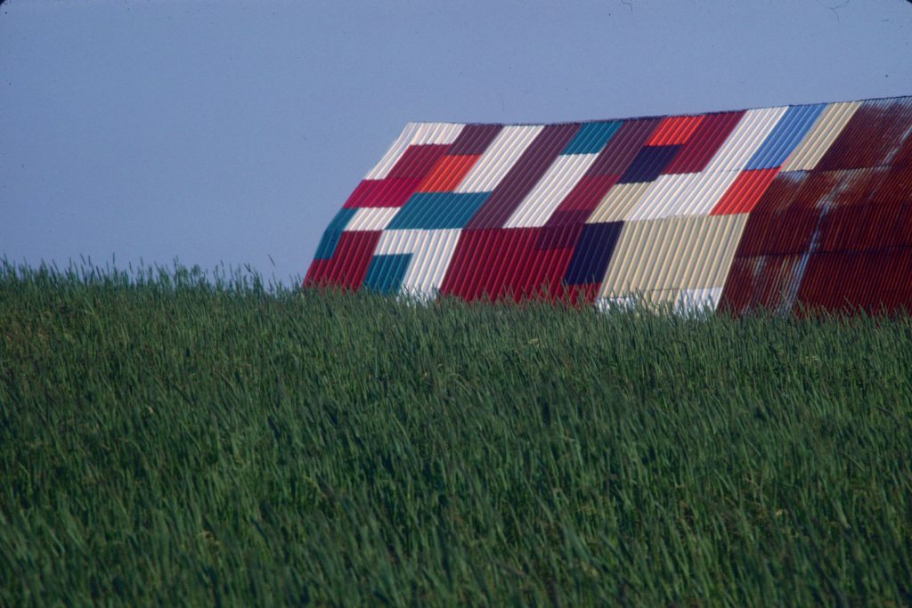 Colour photograph of a green field with in the background the roof of a building in sheet metal with multicolored parts.