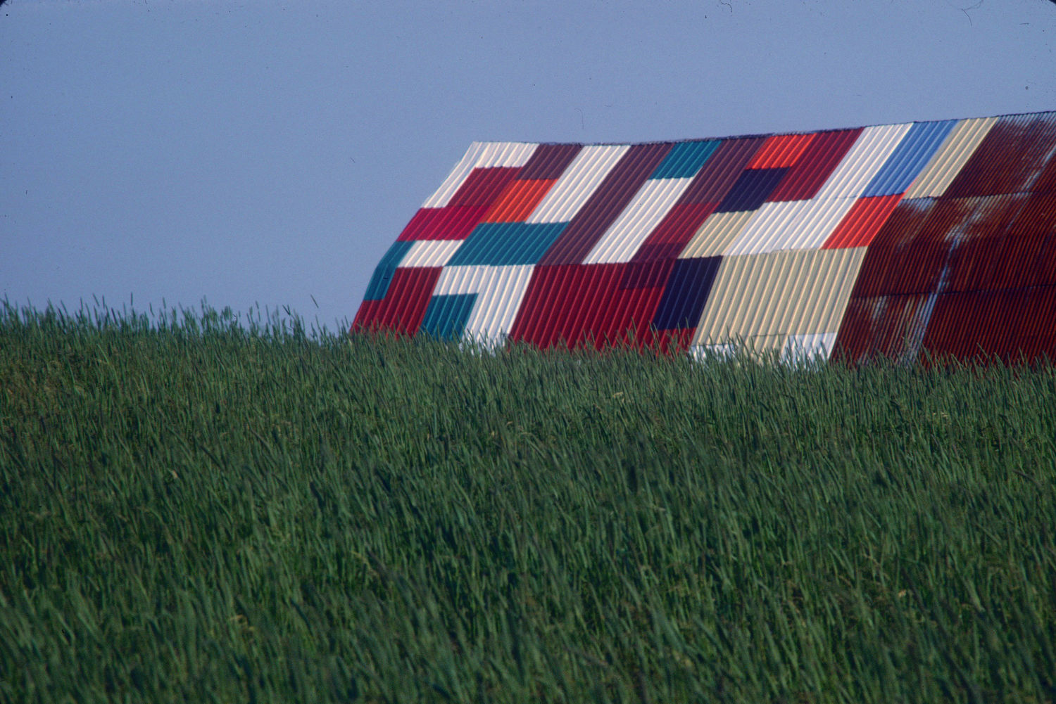 Colour photograph of a green field with in the background the roof of a building in sheet metal with multicolored parts.