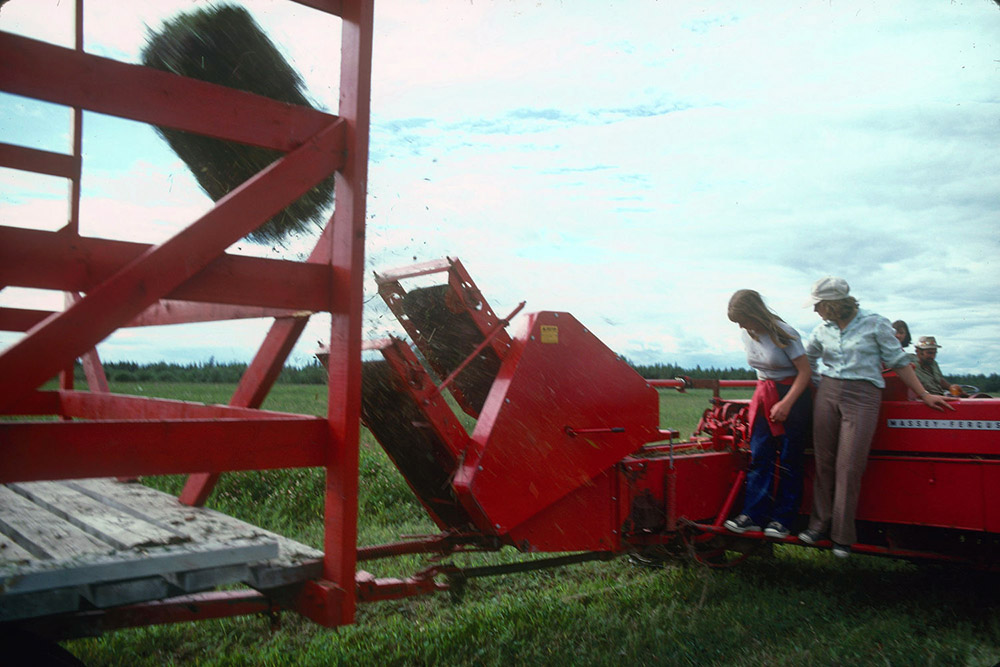 Coloured picture of two people overlooking the production of a hay bale. We see a bale being ejected in a wagon.