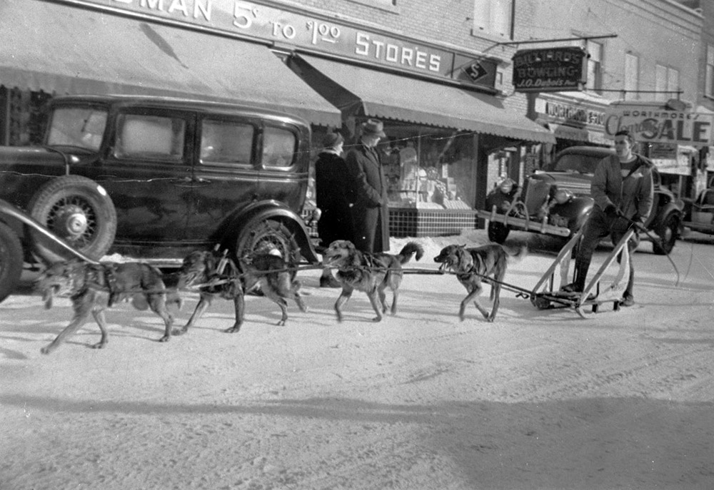 Photographie noir et blanc d'un homme en traîneau tiré par des chiens attelés sur une rue commerciale en ville, en hiver.