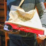 A museum interpreter is holding a red wooden seed planter with a metal rotary distributor and a cotton pouch.