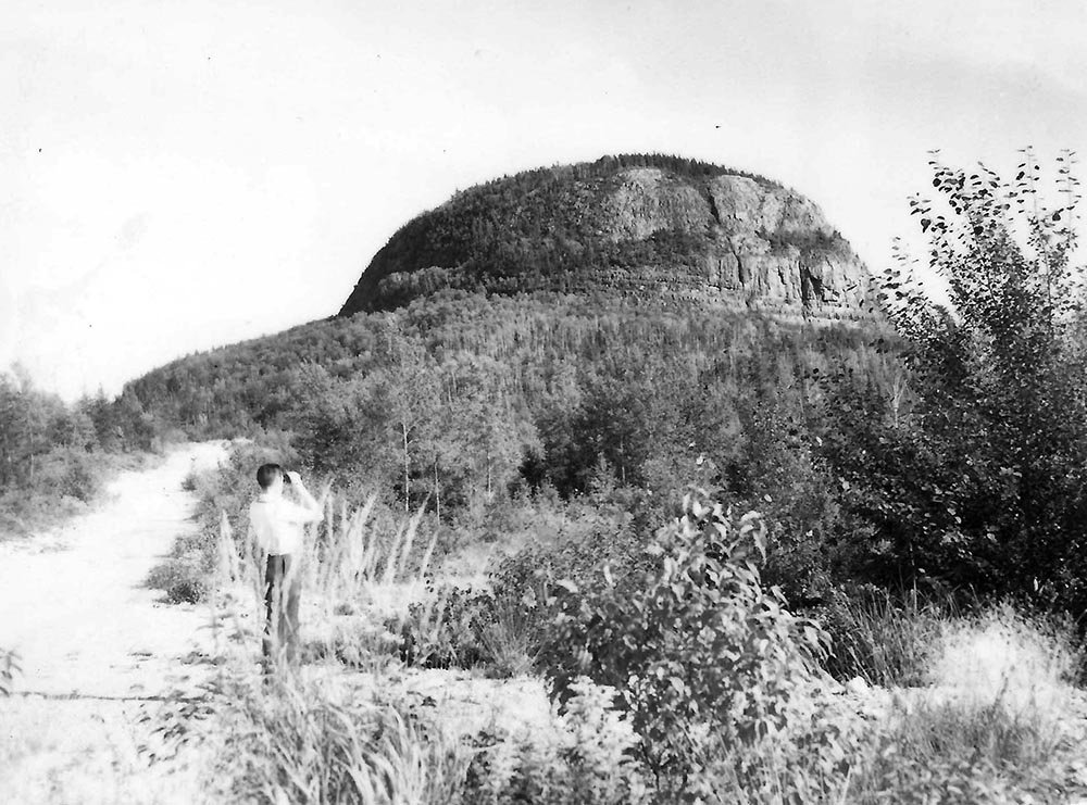 Photographie noir et blanc d'une montagne avec à l'avant-plan un homme vu de dos qui observe à l'aide de jumelles.