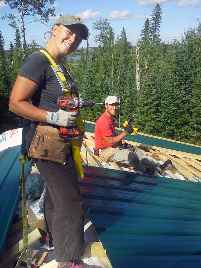 View from the roof of a house in a rural area. Two people are working on the installation of the roof. Large trees and a river in the back.