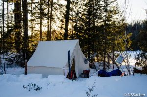 Photograph of a prospector's tent in the forest during the winter. Equipment is lying in front of the tent.