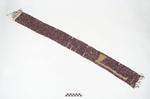 Belt with woven pearls, long rectangular shaped, with purple and white stains. We can see the shape of an axe as decoration.