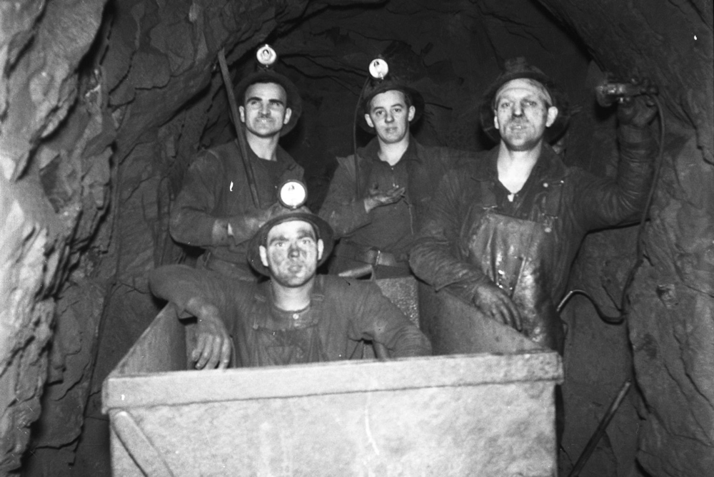 Four miners in working clothes underground, in a gallery posing for the photographer. One of the miners is sitting in a conveyer.