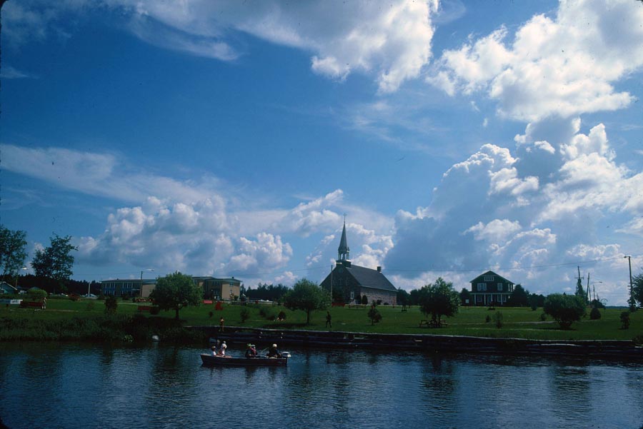Coloured picture of an outside summer view of a village, with a church at the centre. In the forefront, we can see a boat with people on a lake.