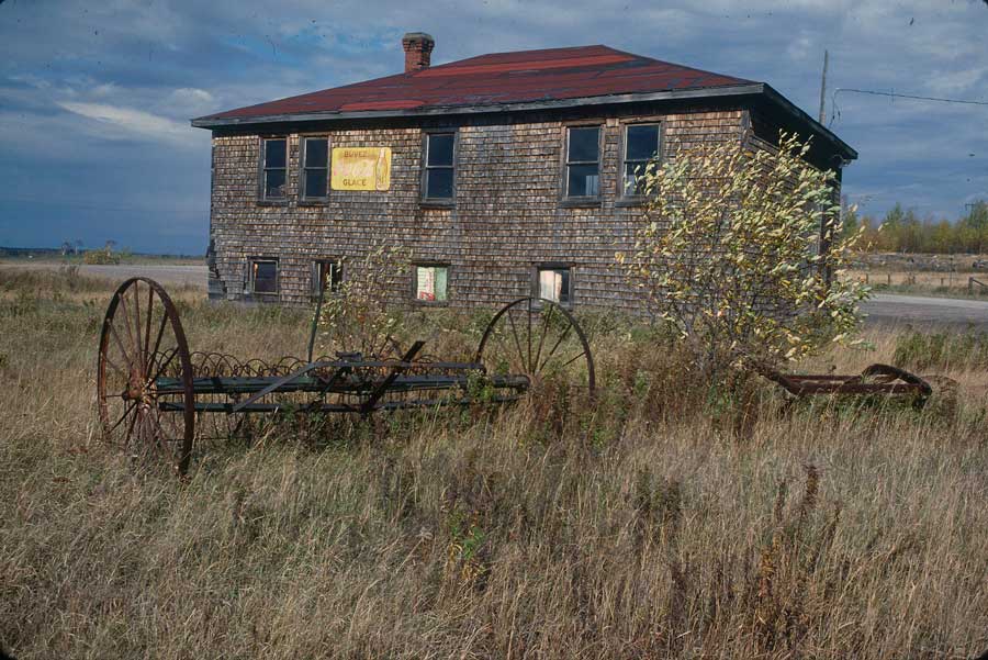 Coloured picture of a wooden building with abandoned agricultural equipment.