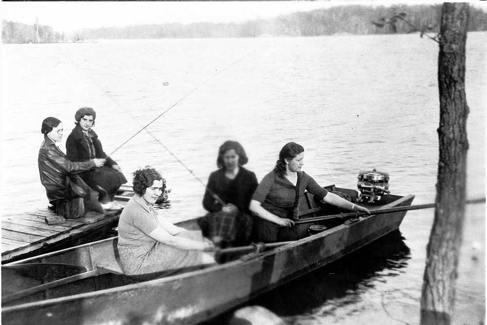 Black and white picture of women on a fishing trip, fishing rods in hand, some on a dock and some on a canoe.