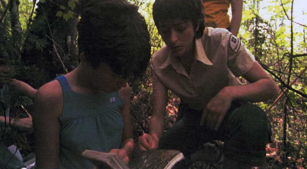 Photographie couleur de deux personnes accroupies en forêt, qui regardent un cahier. L'une d'entre elles porte un costume d'employé de parc national québécois.