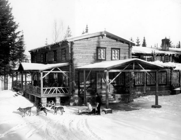 Black and white photo of Auberge La Sapinière in winter 1937; a dogsled is standing in front of the log-built lodge.