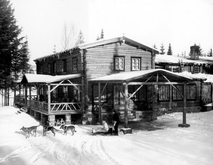 Black and white photo of Auberge La Sapinière in winter 1937; a dogsled is standing in front of the log-built lodge.