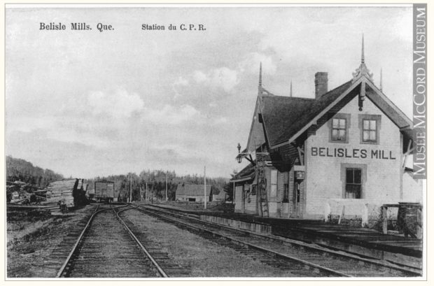 Vintage postcard showing the Bélisle’s Mills Railway Station; apart from the building, the railway tracks can be seen and stacks of wood ready for loading.