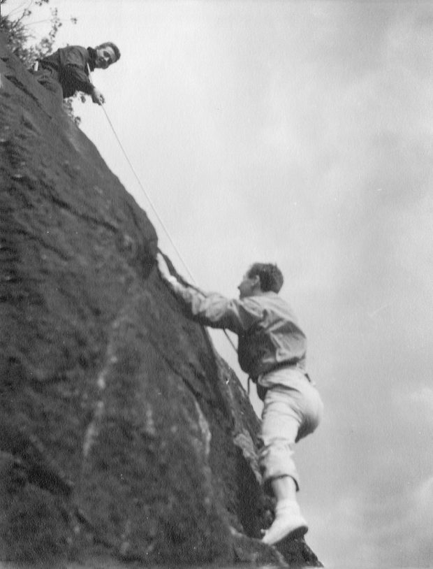 Two climbers, one at the top of the rock face holding a rope by means of which the second climber is still ascending.