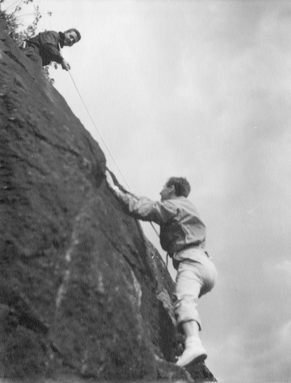 Two climbers, one at the top of the rock face holding a rope by means of which the second climber is still ascending.
