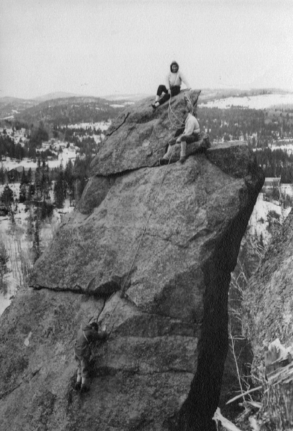 Three climbers in winter, two on the rocky summit and a third still climbing up, against a background of snowbound countryside.