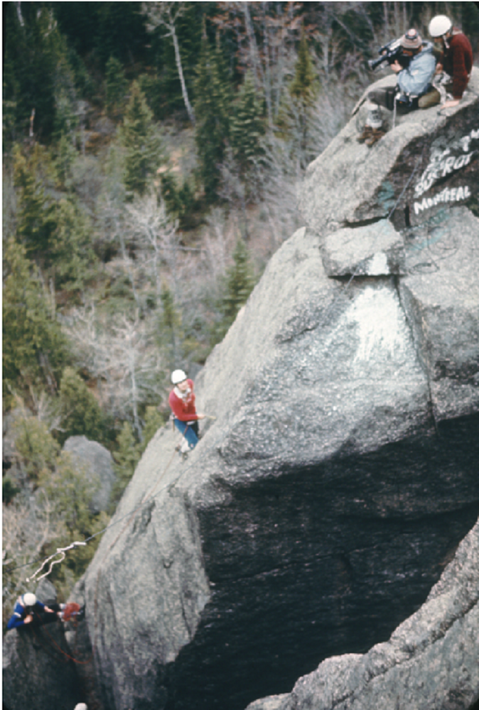 A group of climbers on a rock face high in the forest. A cameraman and a belayer are at the summit; a woman is ascending the rock face.