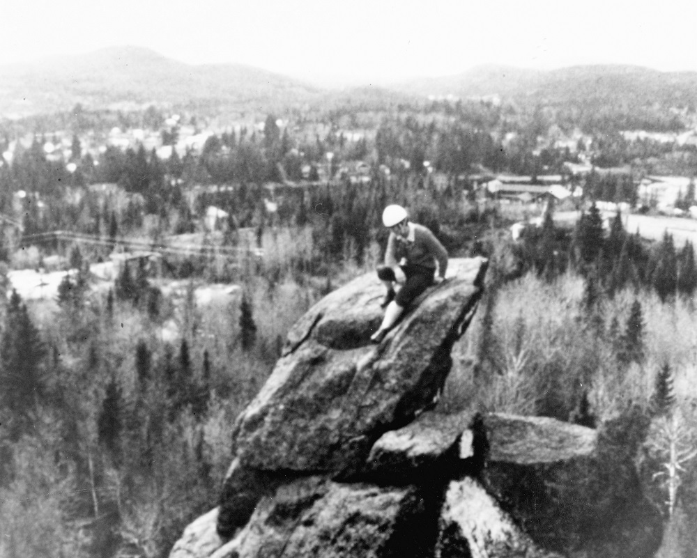 A woman sits on the top of a high rock. In the background is the countryside of Val-David.