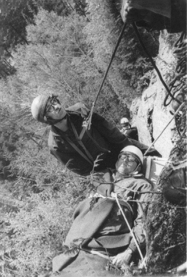 View from above of two men on a rock face in mid-descent; one is strapped to a stretcher and the other is assisting the descent by means of ropes.