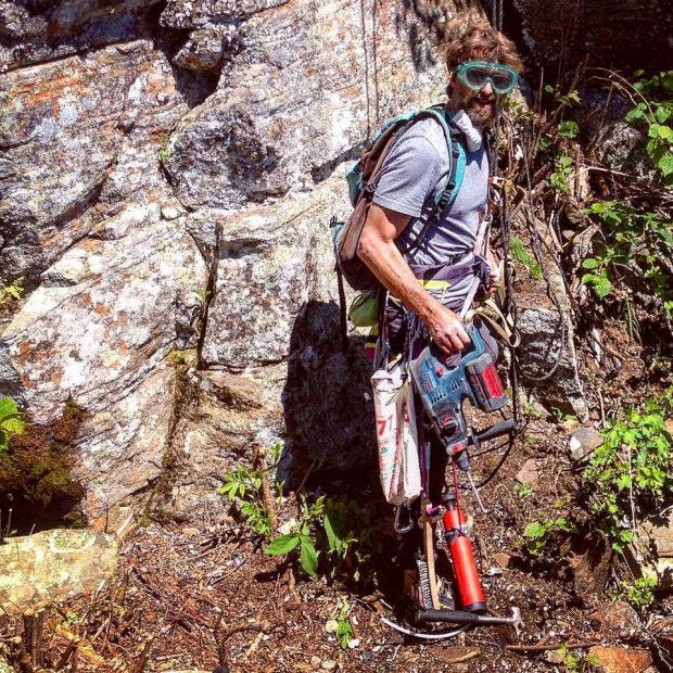 Close-up of a climber wearing safety glasses, brushes and other items attached to the harness around his waist. He is also holding a percussion drill in his hand.