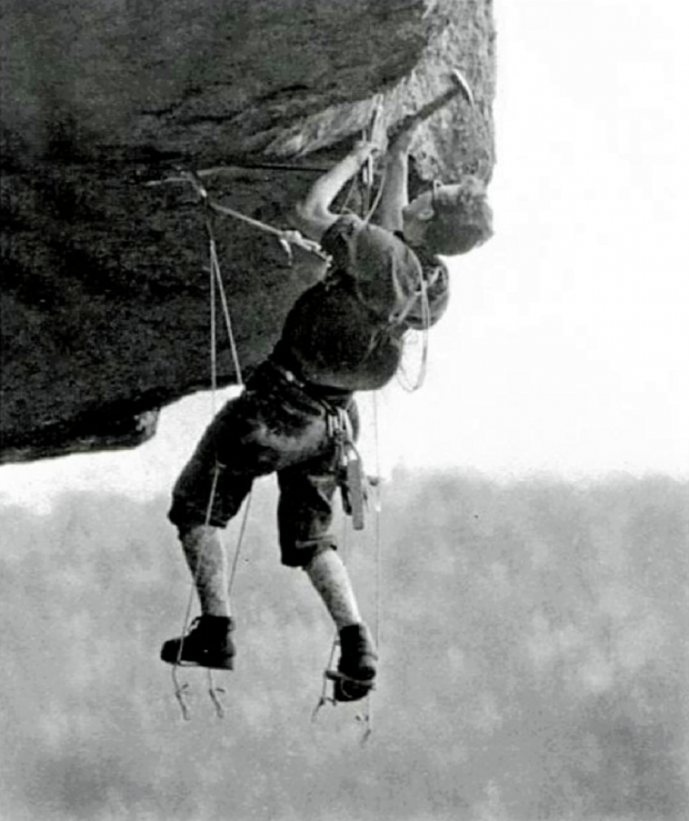 A vintage photo of a climber suspended in mid-air, supported by straps and a short ladder, who is hammering in a piton against a background of sky and the treetops.