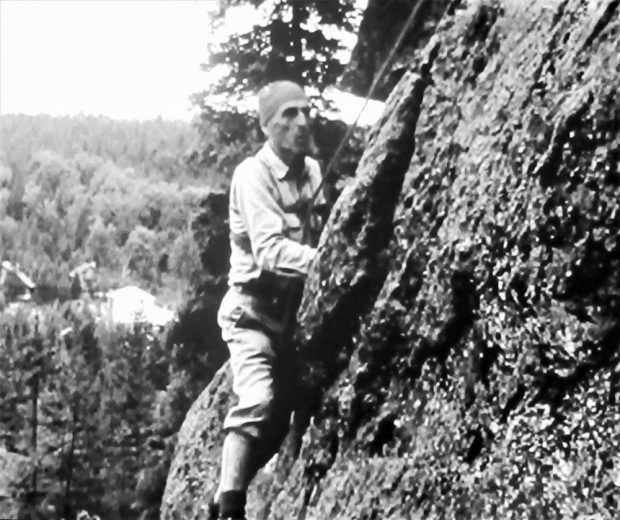 Vintage photo of a climber hanging onto a rock face with hands and feet, against a background of sky and treetops.