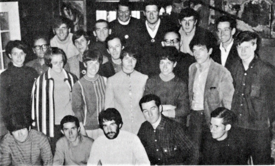 Photo en noir et blanc plein cadre d’une vingtaine de personnes réunis dans une salle.