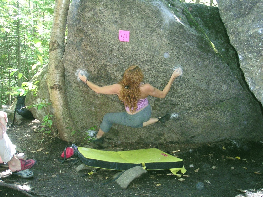 Close-up of a young woman hanging on with hands and feet on a boulder only centimetres from the ground, where her crash pad can be seen below.
