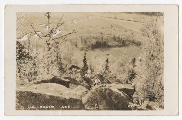 Vintage postcard looking down from a summit; two men are climbing up over a series of boulders. In the background is part of the valley of Val-David.