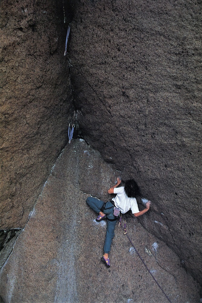 View of a climber on a brownish-orange rockface in a position demonstrating her lower-body and leg flexibility.