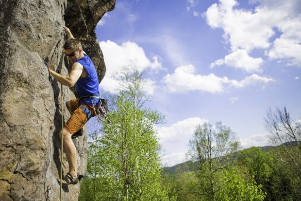 Close-up in colour of a climber hanging on with hands and feet against a background of blue sky, a few clouds and the treetops.