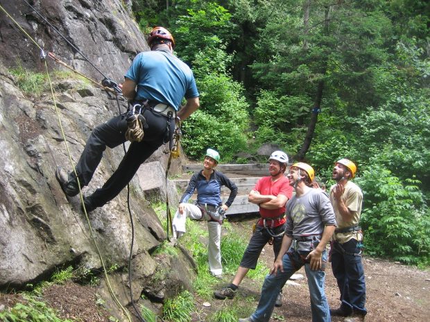 Five people; one man attached to a rock face at a low height; three men and a woman observing from the ground.