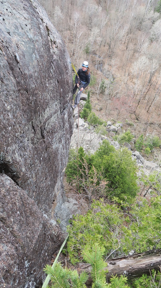 Photo of a young woman attached to a yellow rope on a rock face, against a background of the forest in late fall.